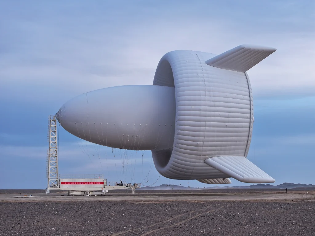 Large helium filled tethered airborne wind turbine prototype on a test site, showing a floating aerostat with stabilising fins connected to the ground by multiple cables against an open sky.