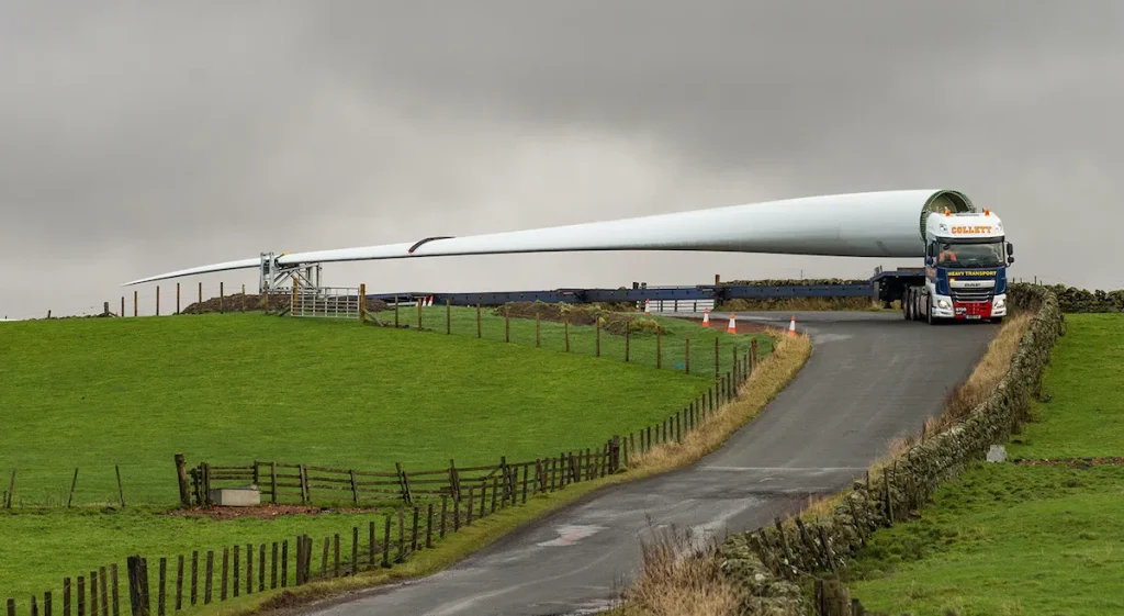 Long wind turbine blade carried on a heavy transport lorry navigating a narrow countryside road bordered by fields and stone walls under overcast skies.
